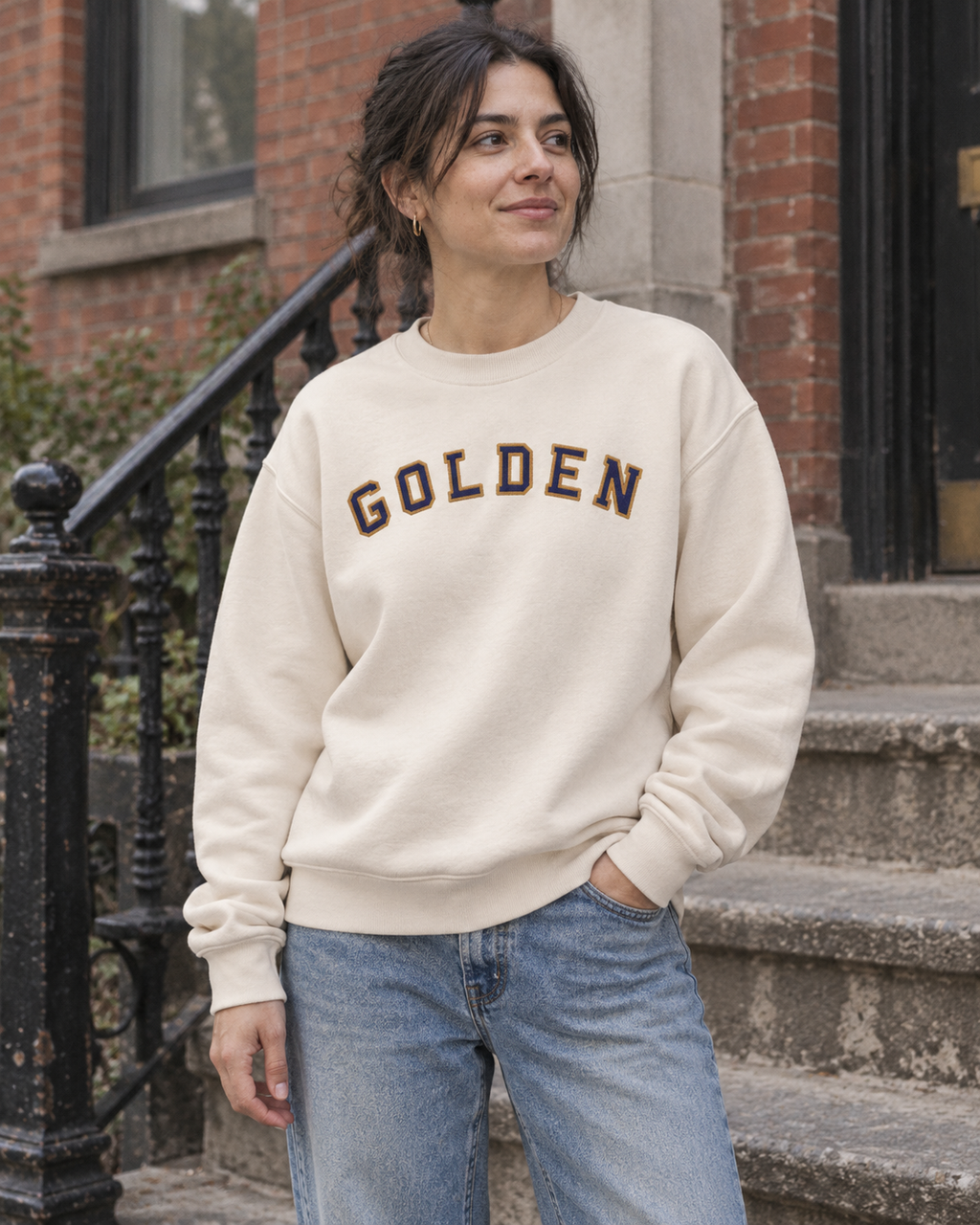 Woman wearing the GOLDEN Varsity Relaxed Crew beside a brick stoop — lifestyle mockup