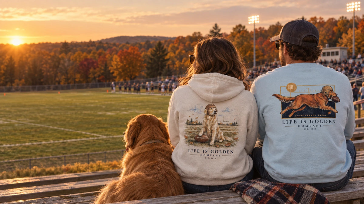 Two football fans wearing The Goal Line crewneck and The Friday Night Lights hoodie, sitting with their golden retriever at an autumn game