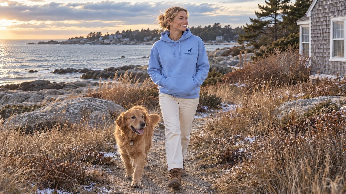 A woman in the Carolina blue Life Is Golden hoodie walking with a golden retriever along a frosty coastal path.