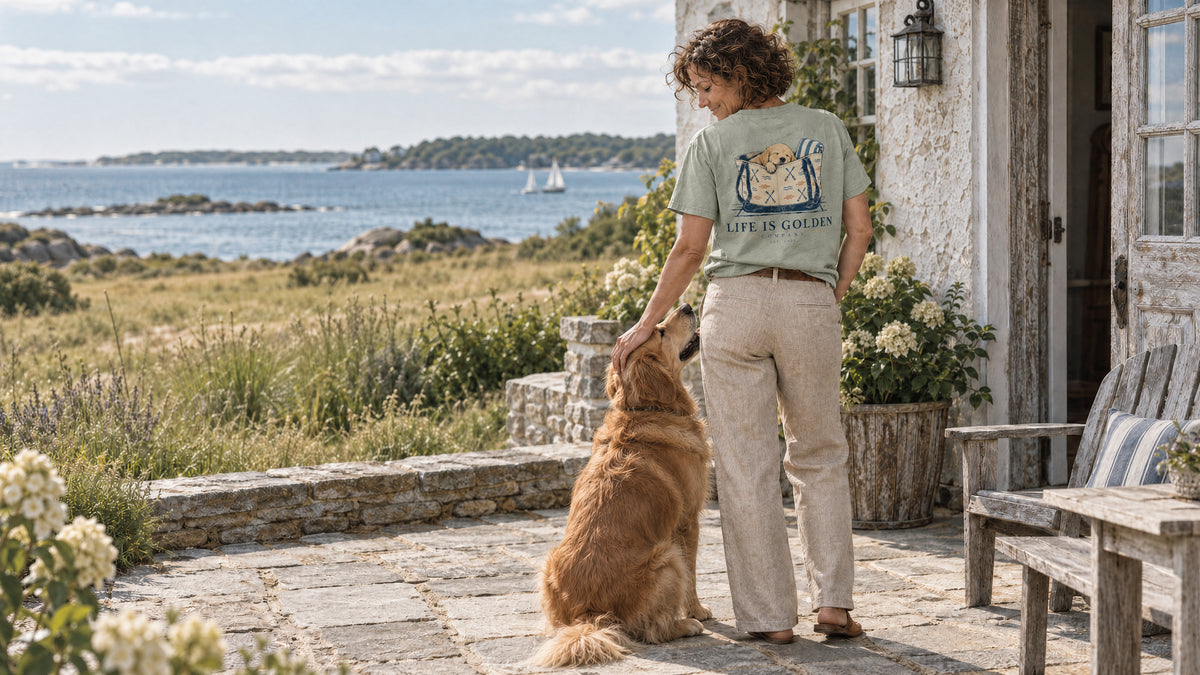A woman in the Bay Beach Bag Nap tee and linen trousers with a golden retriever on a coastal cottage terrace